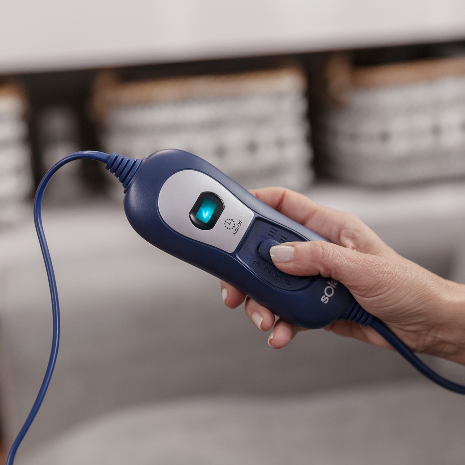 Una mano sostiene un control remoto azul con pantalla iluminada y cable en una habitación.