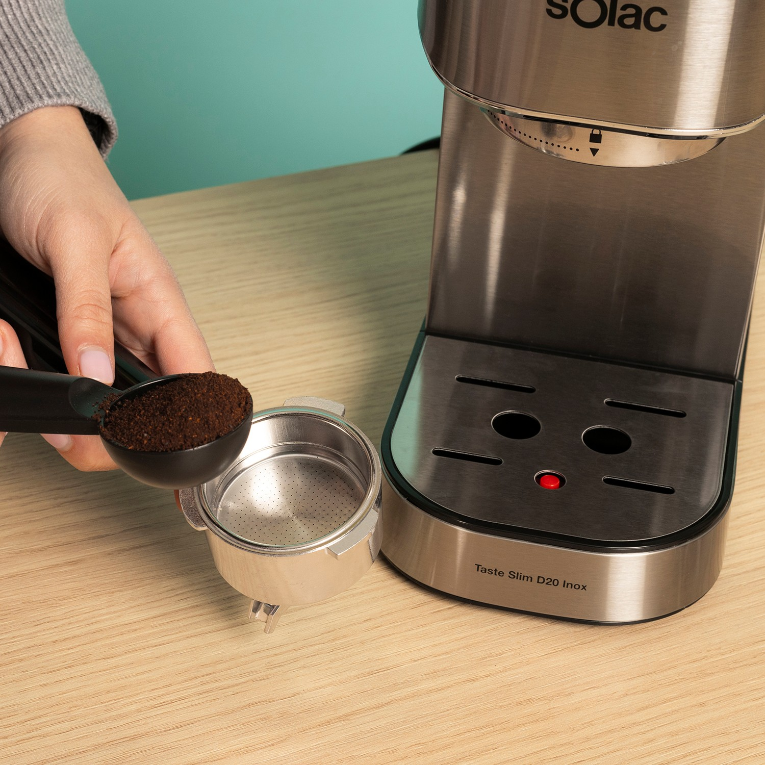 Una mano sostiene una cuchara con café molido sobre un portafiltro junto a una cafetera.