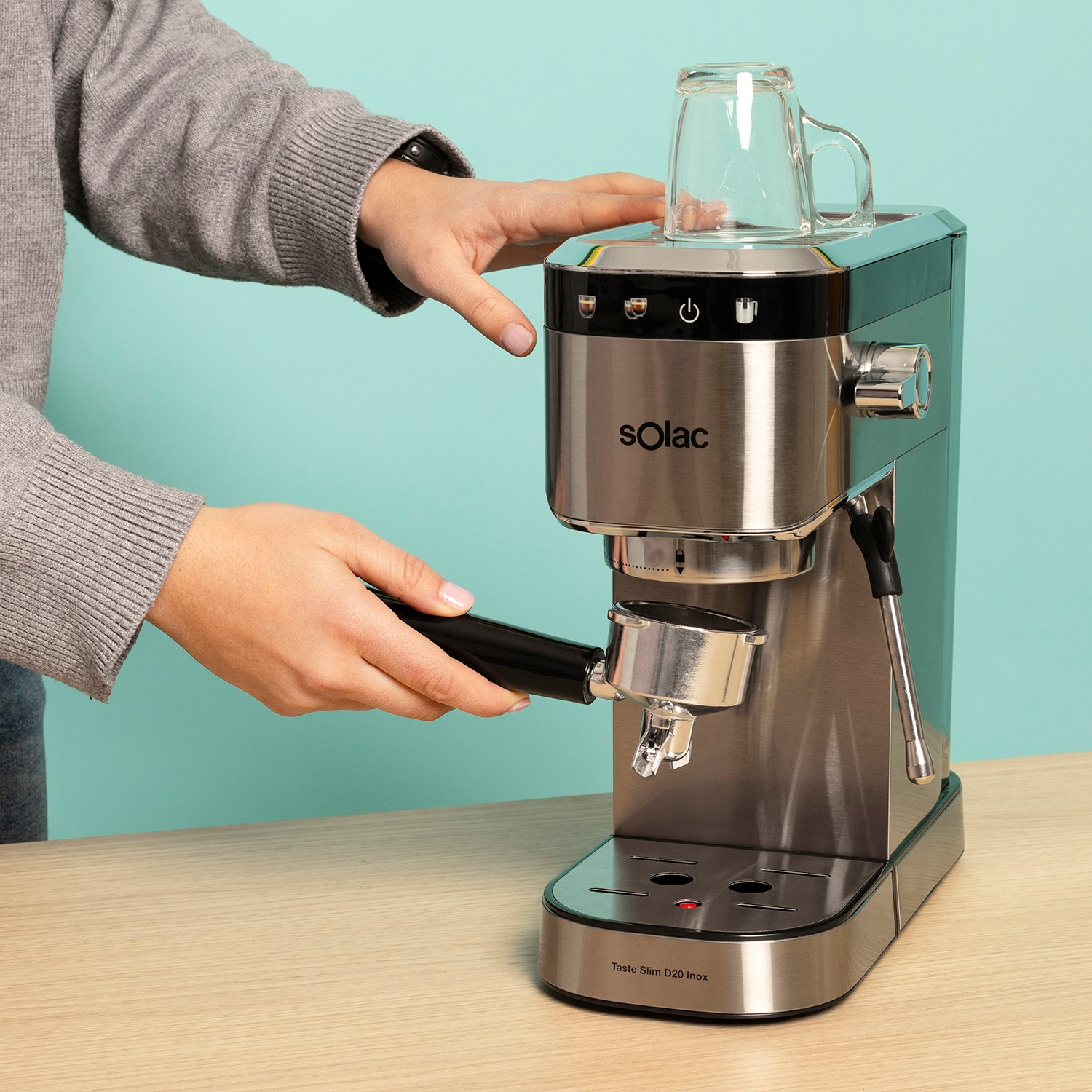 Una persona con suéter gris usa una cafetera Solac de acero inoxidable con un vaso encima.