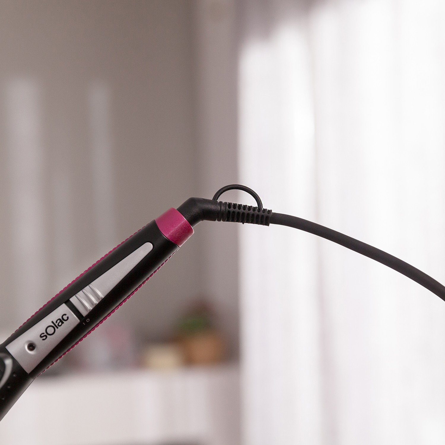 Una plancha de pelo Solac negra con detalles en rosa y un cable de alimentación sobre fondo difuminado.