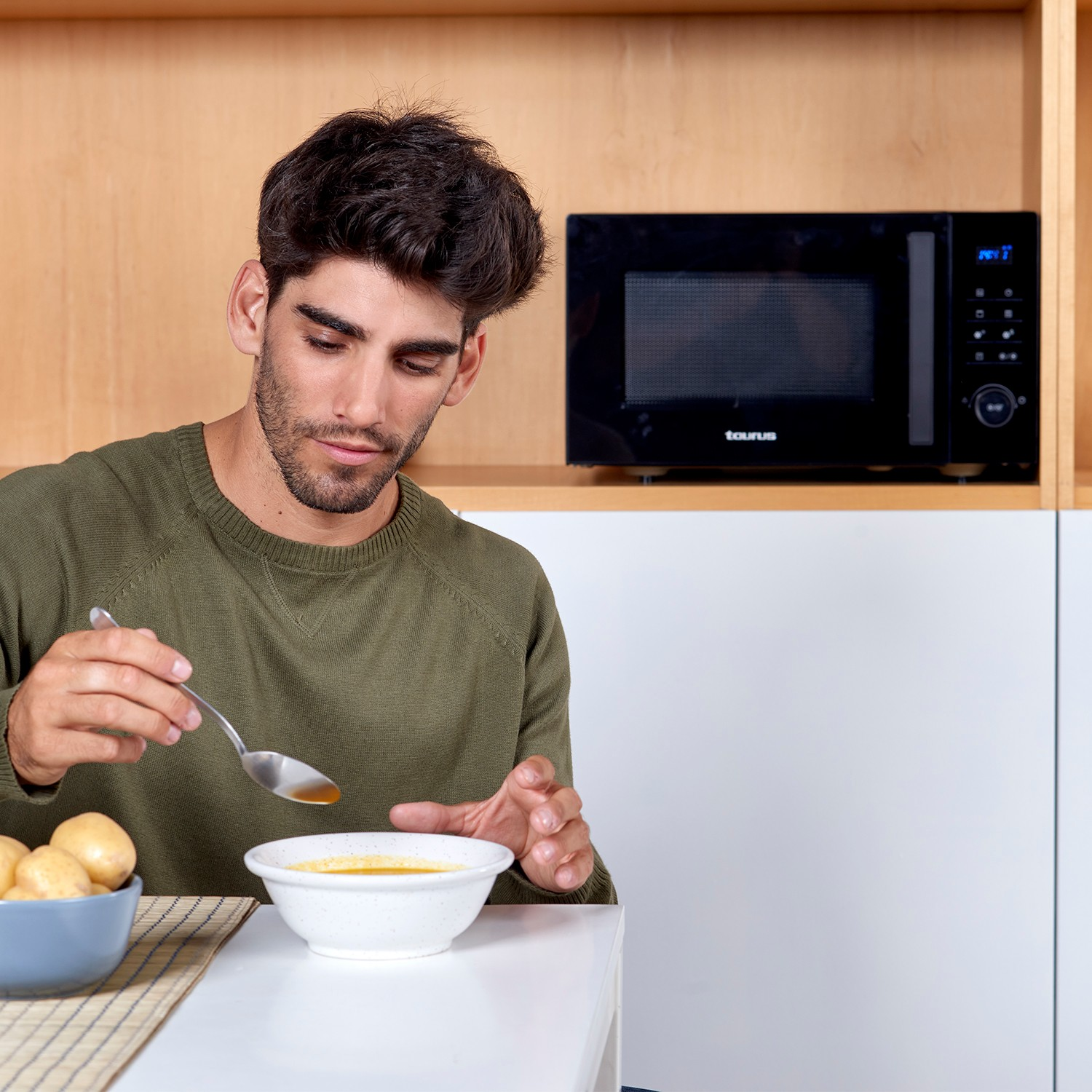 Un hombre con suéter verde come sopa de un bol blanco en una cocina moderna con microondas.