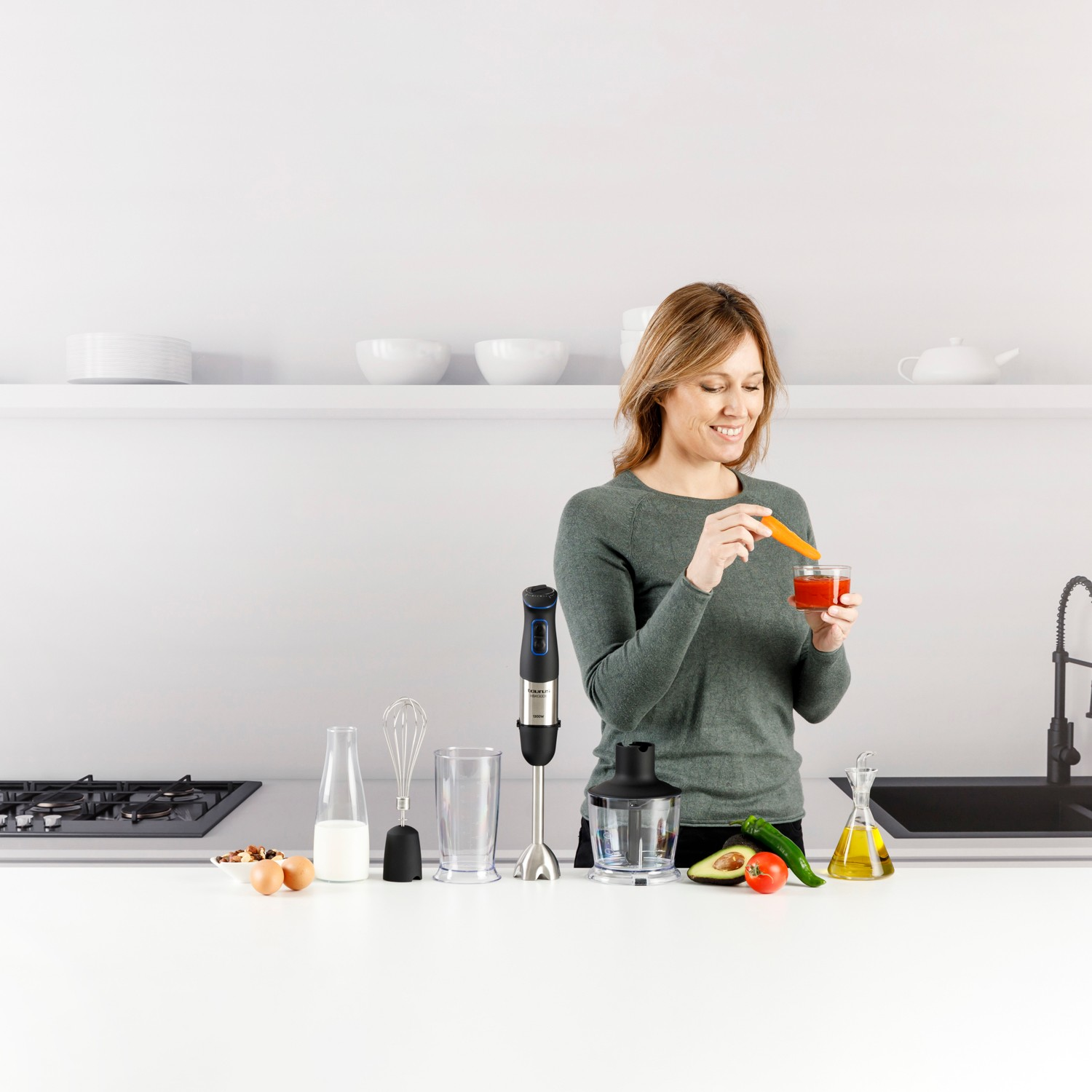 Mujer en la cocina sosteniendo zanahoria y vaso de salsa junto a batidora de mano, ingredientes y accesorios.