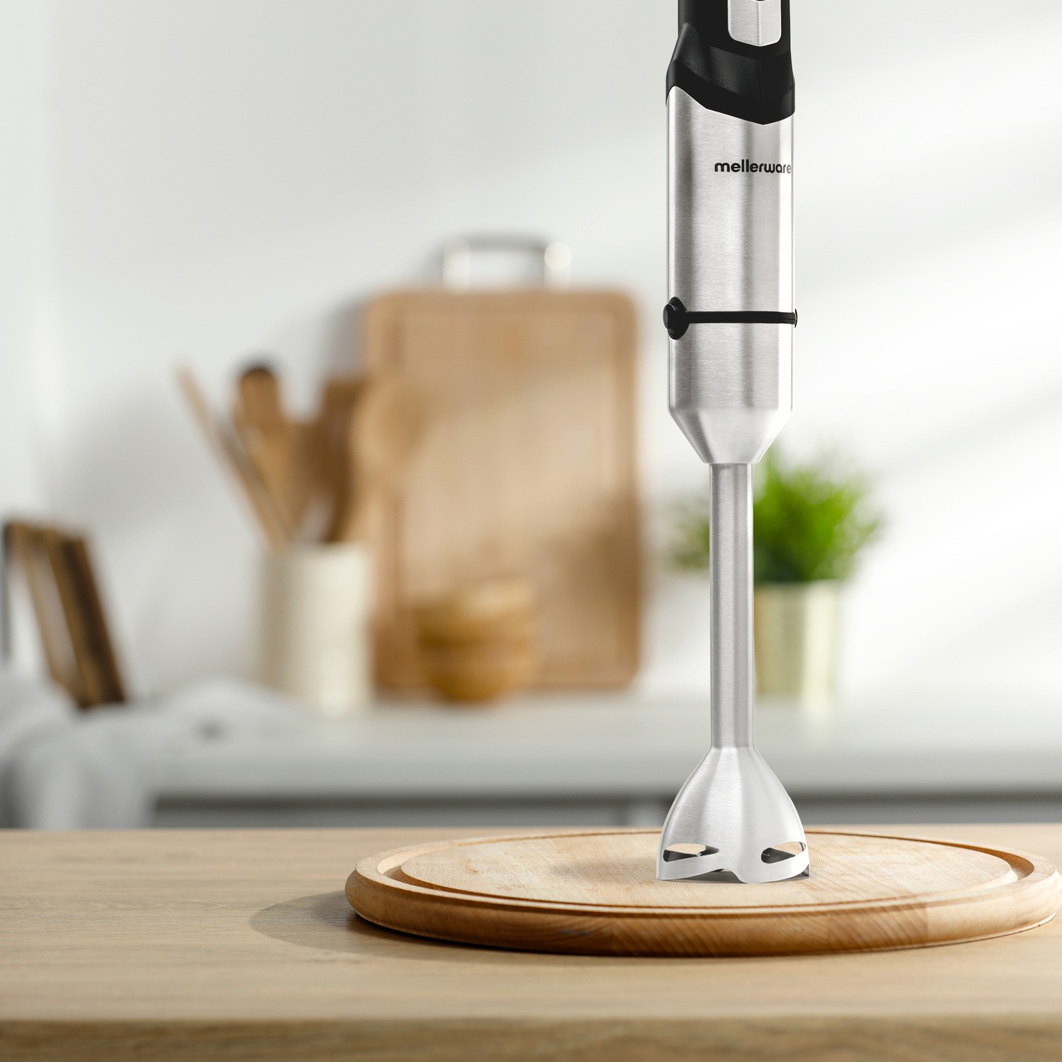 Una batidora de mano plateada se alza sobre una tabla de cortar de madera redonda en una cocina.