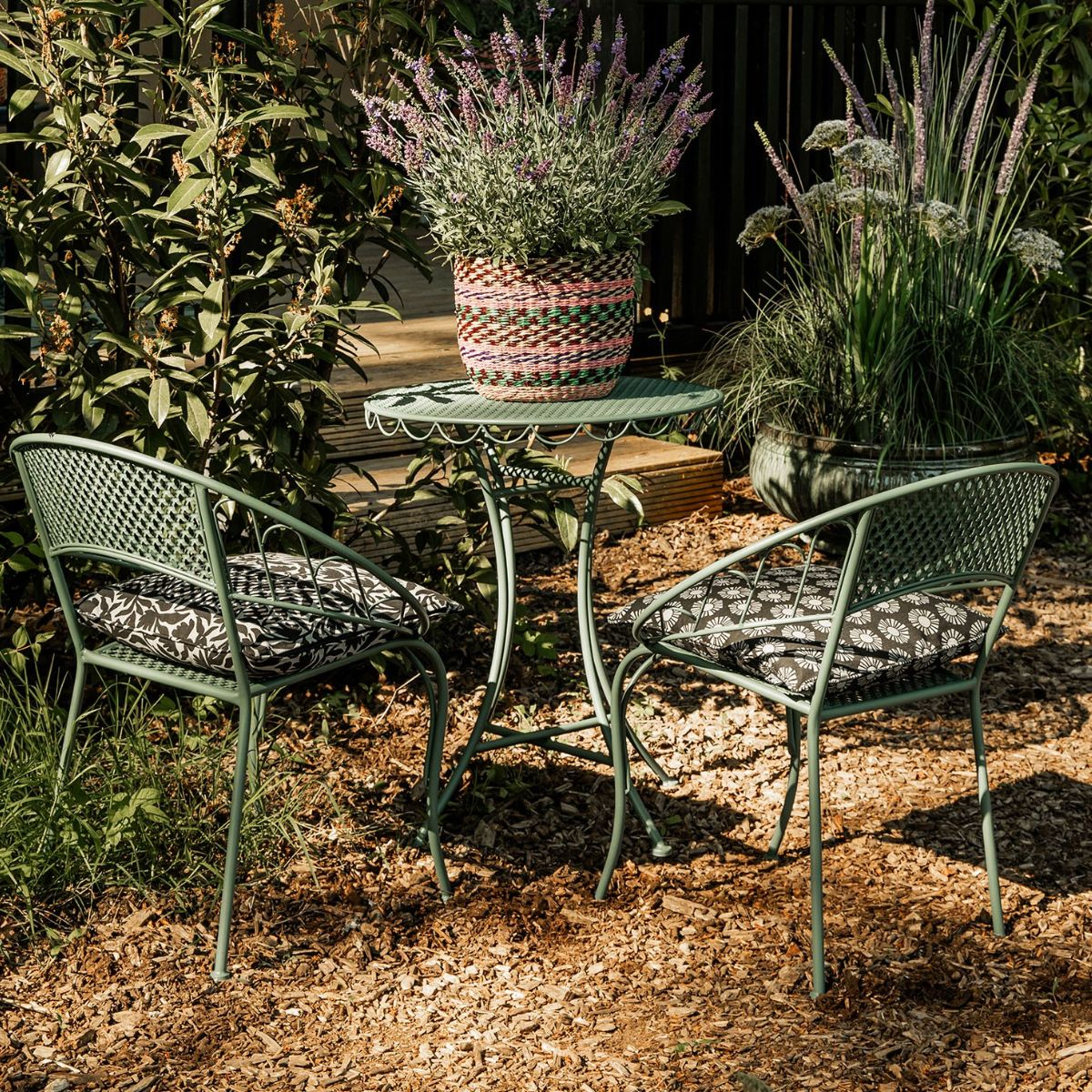 Grüner Gartentisch mit zwei Stühlen und Kissen steht im Garten vor Pflanzen.