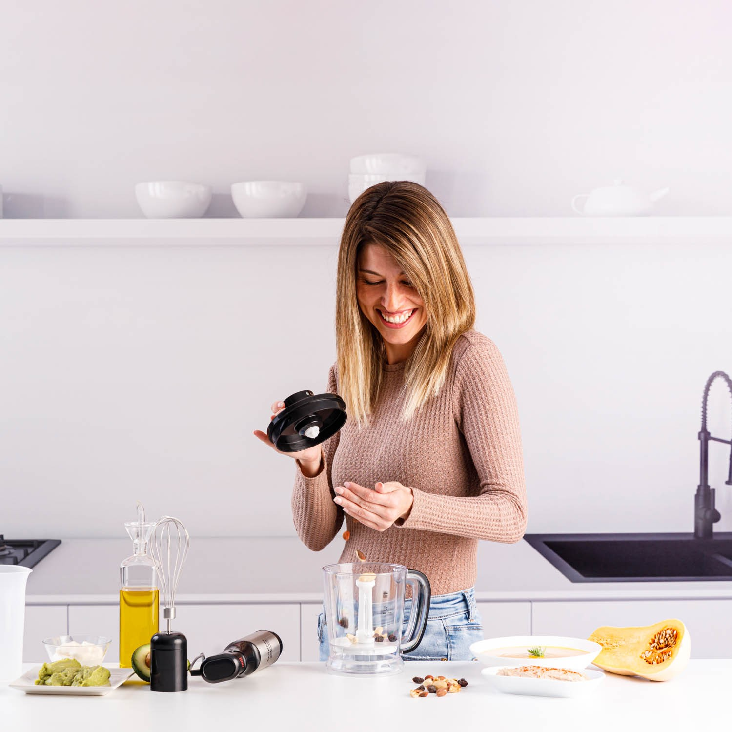 Una mujer sonríe mientras echa frutos secos en un procesador de alimentos en una cocina luminosa.
