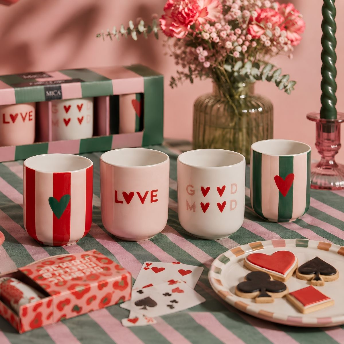 Four patterned mugs, flowers in a vase, playing cards, and heart cookies on a striped table.