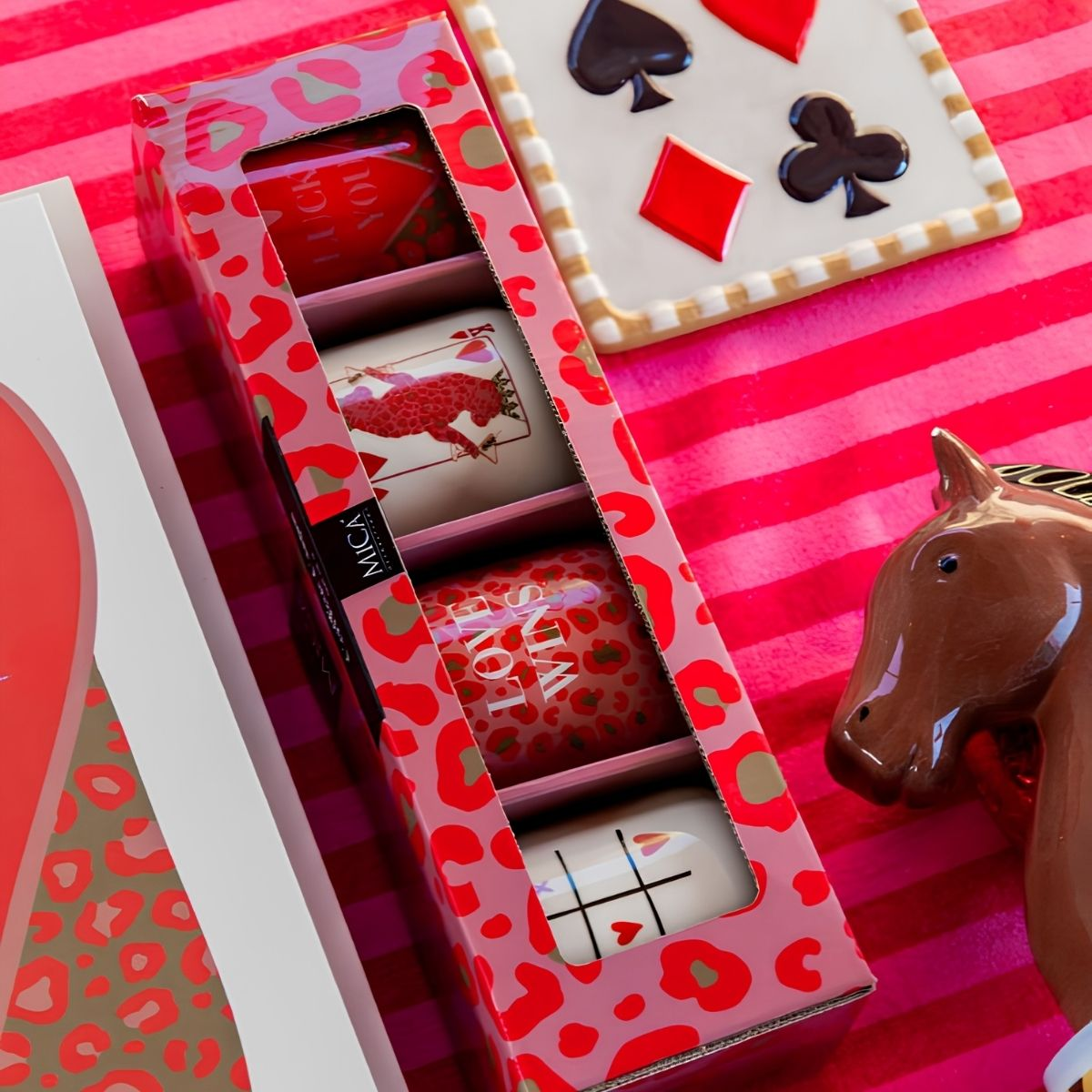 A pink gift box with four mugs, a playing-card design tray, and a brown horse figurine on a red striped surface.