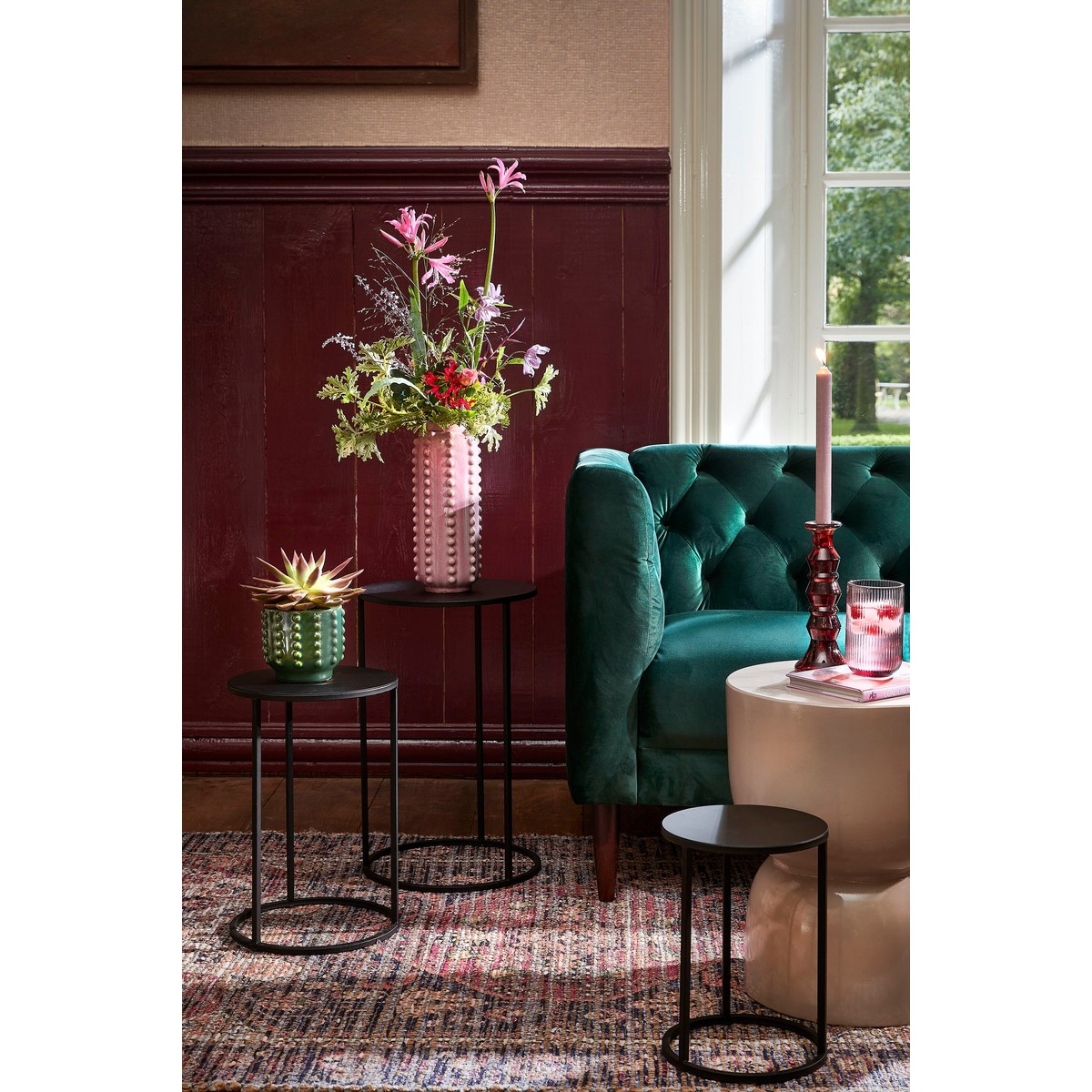 A dark green tufted sofa sits next to small black tables and a pink vase with flowers.