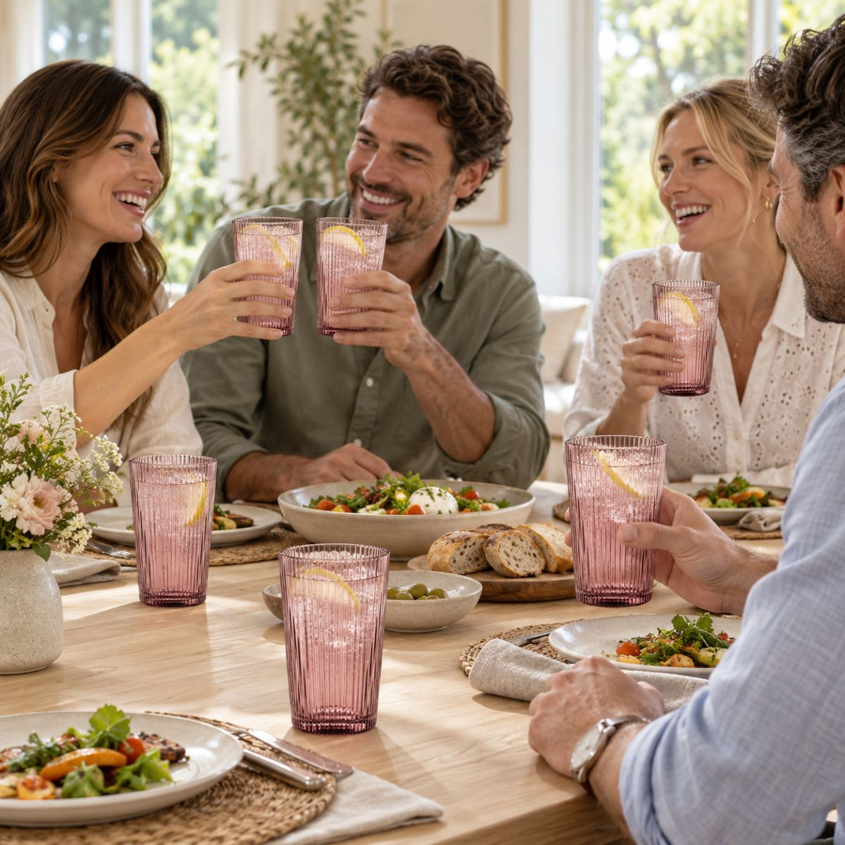 Four friends laughing and toasting with pink glasses of lemonade and food at a wooden table.