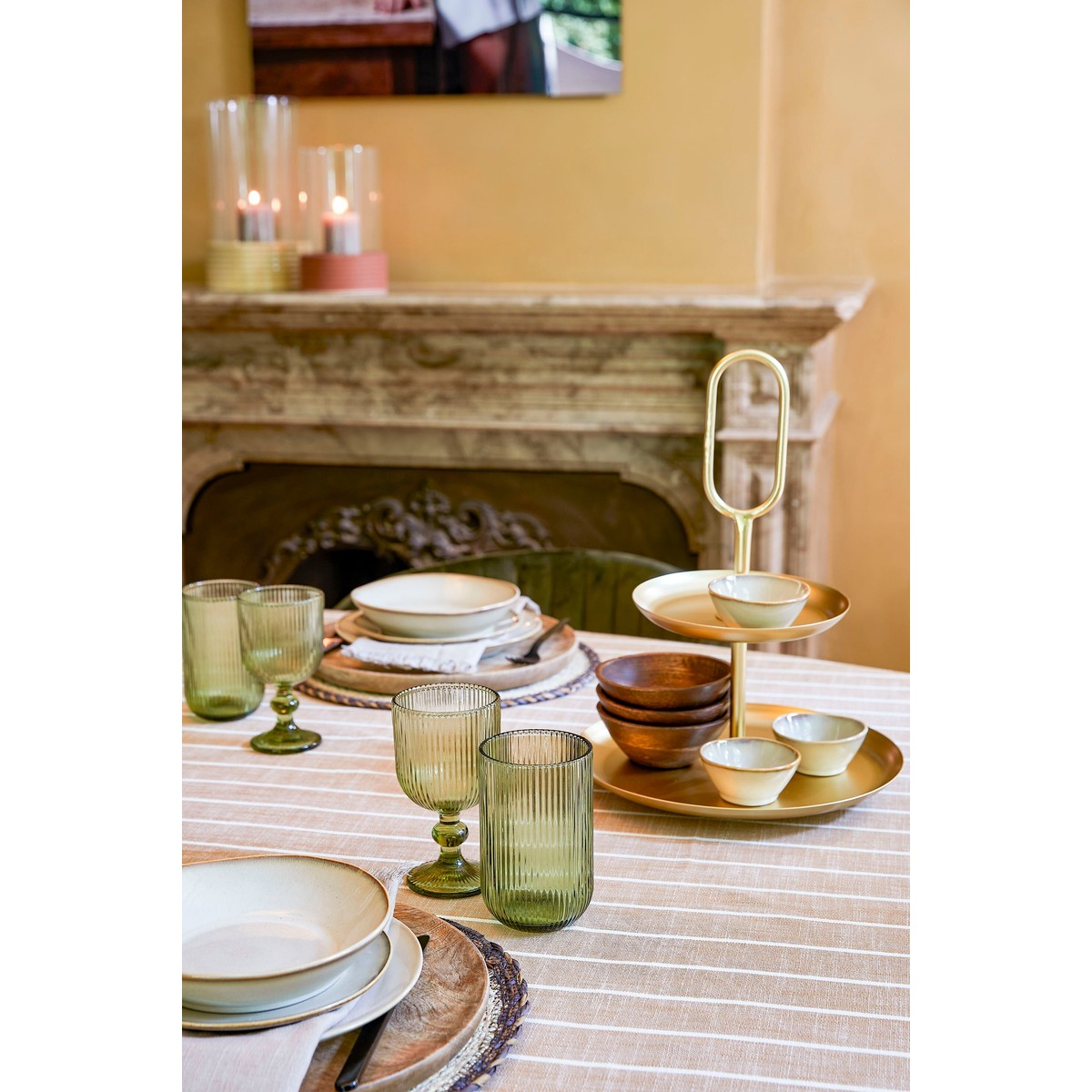 Set table with green glasses, plates, and a gold tiered stand in front of a fireplace.