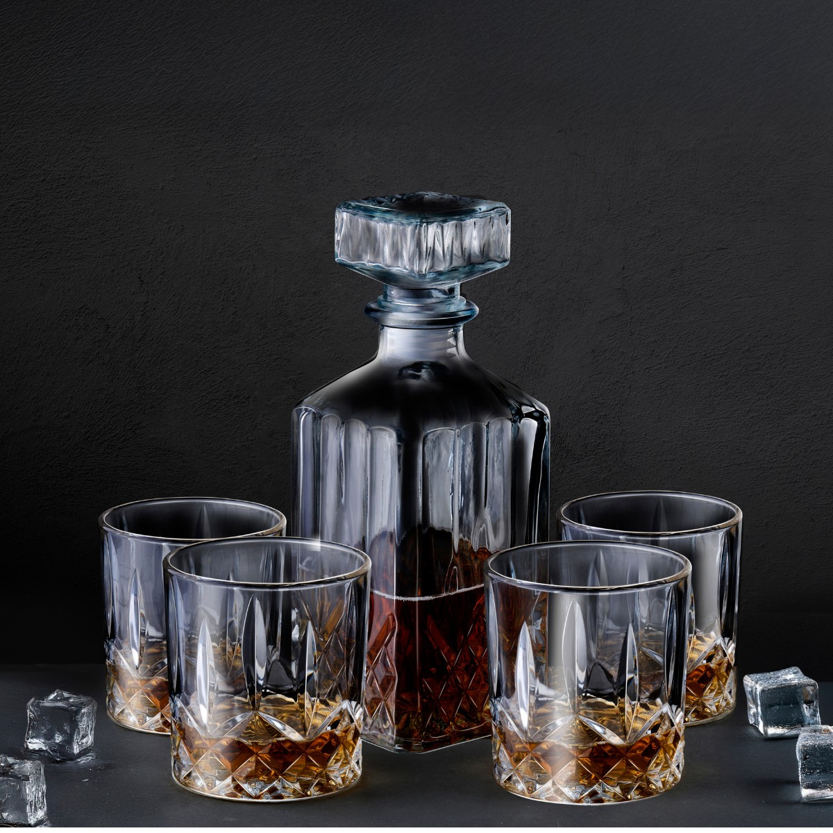 A whiskey decanter and four glasses with whiskey and ice cubes on a dark background.