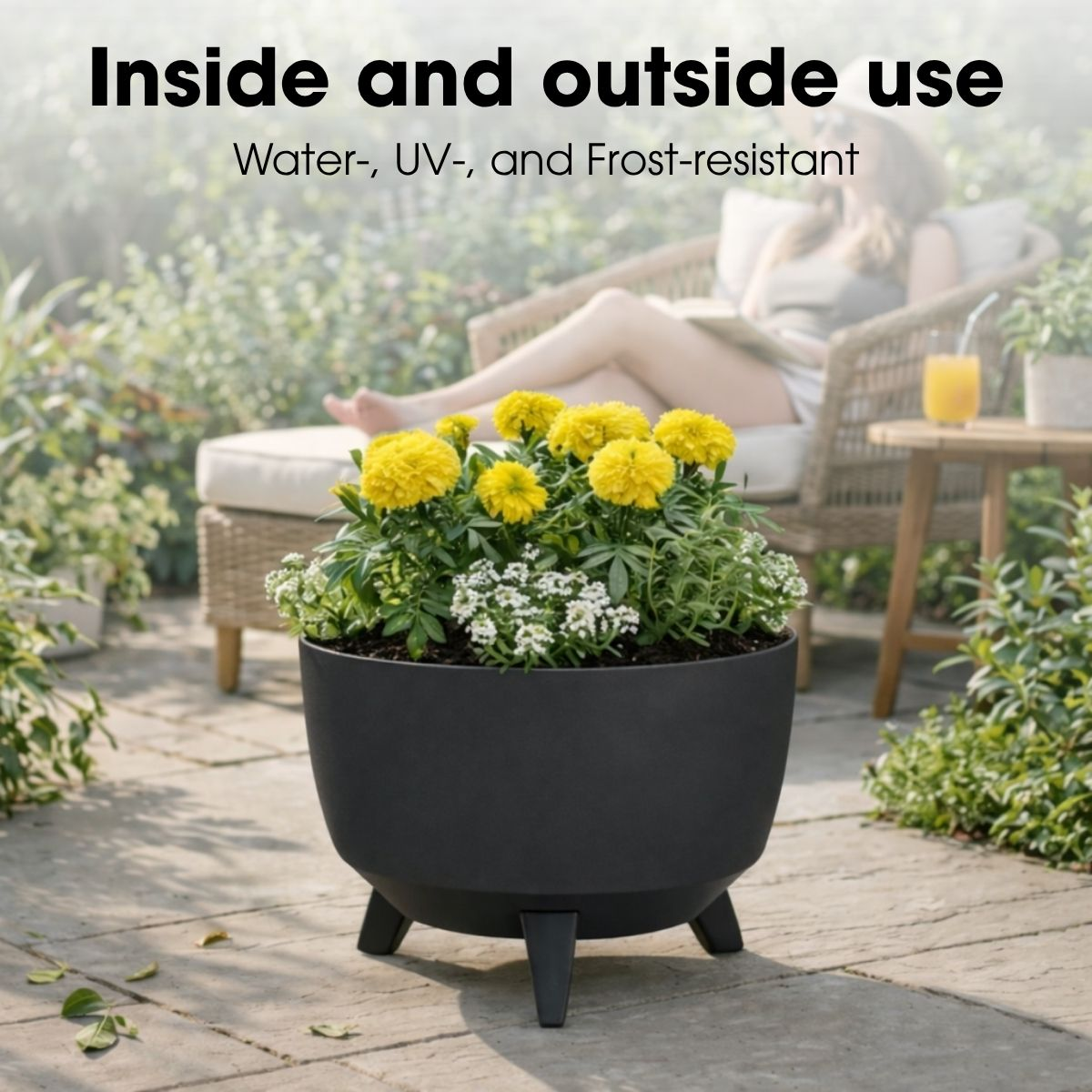 Une jardinière noire avec des fleurs jaunes et blanches est sur un patio, une femme se reposant en arrière-plan.