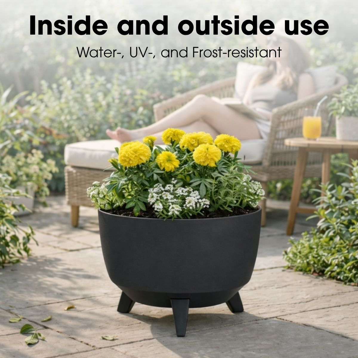 Un pot de fleurs rond noir avec des fleurs jaunes et blanches sur une terrasse, avec une femme au repos en arrière-plan.