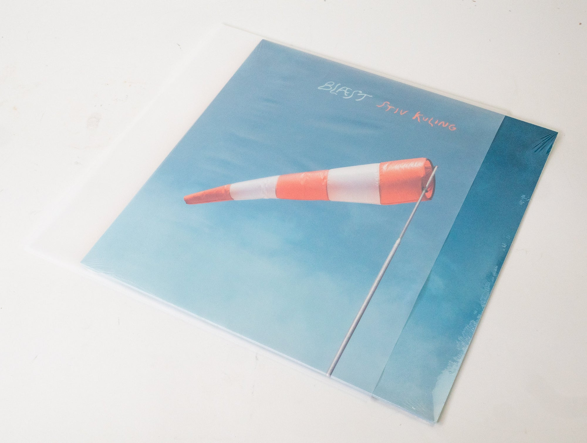 Eine Schallplattenhülle auf einem weißem Tisch zeigt einen orange-weißen Windsack vor blauem Himmel mit Text.
