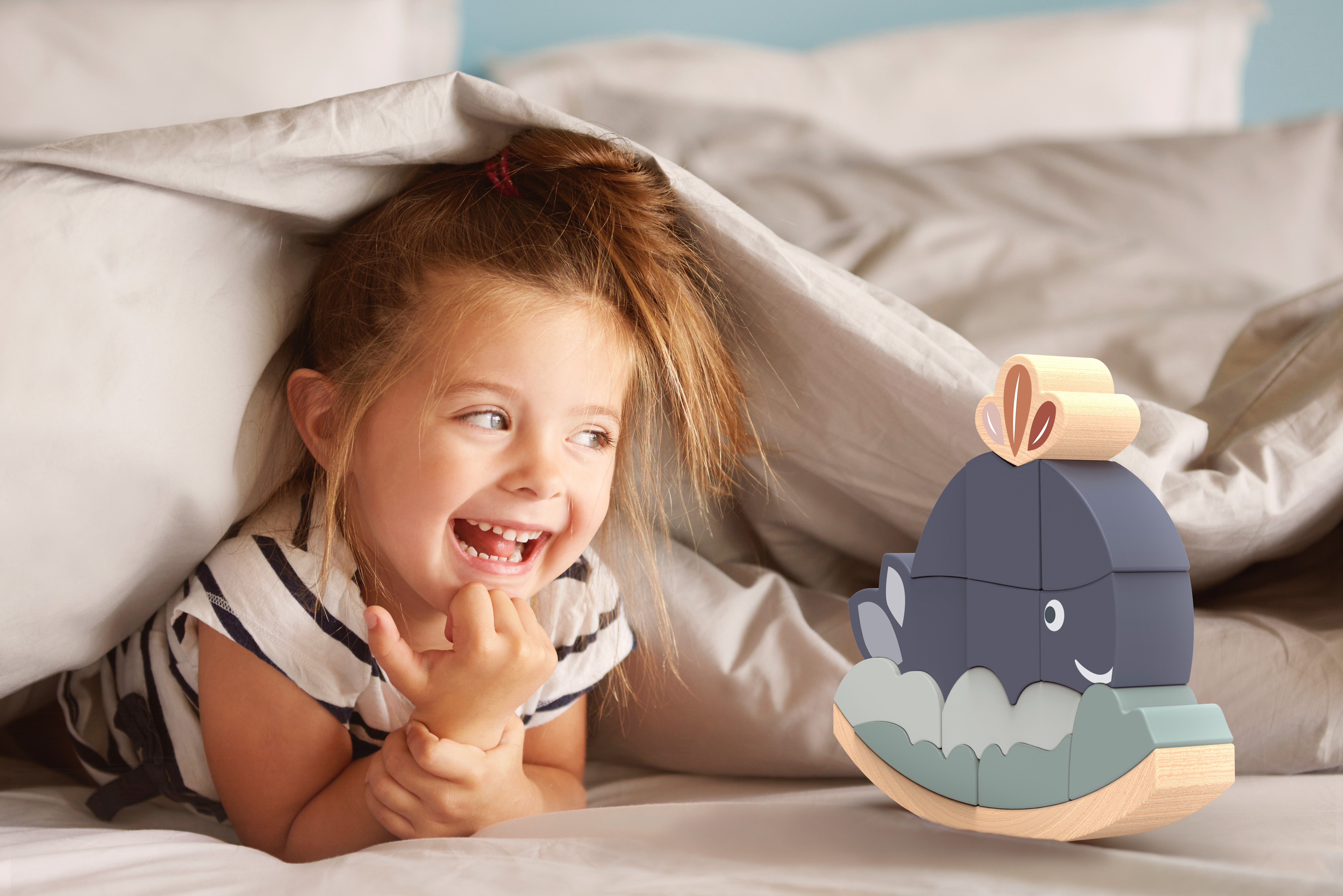 Ein kleines Mädchen lacht im Bett unter einer Decke neben einem Holzspielzeug.