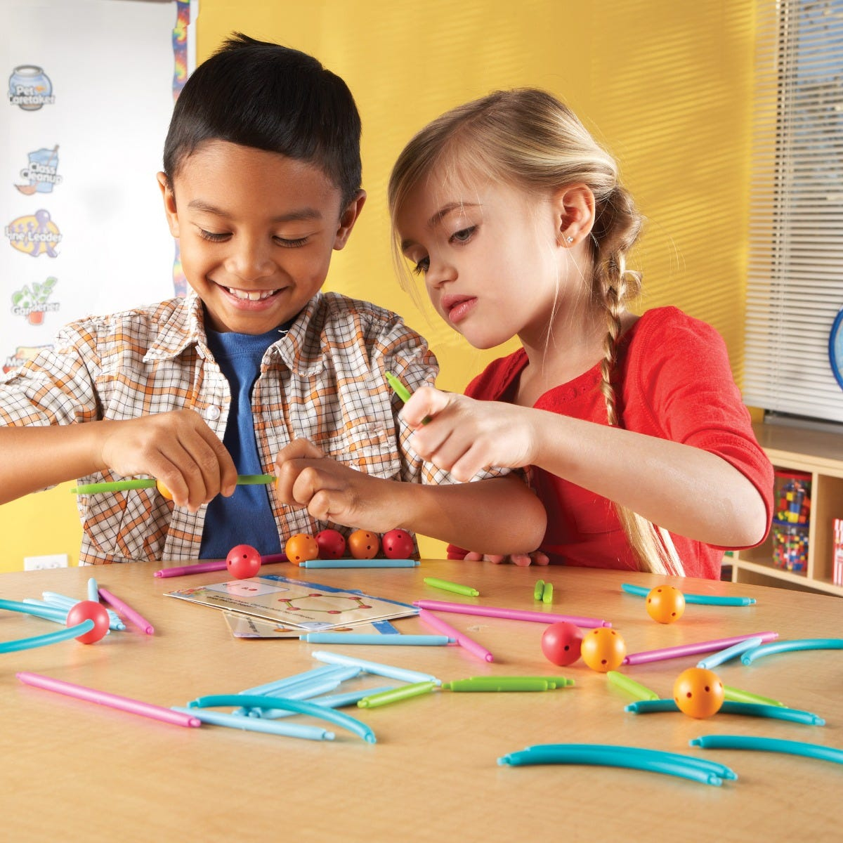 Zwei Kinder lächeln und spielen an einem Tisch mit bunten Konstruktionsstäben und Kugeln.