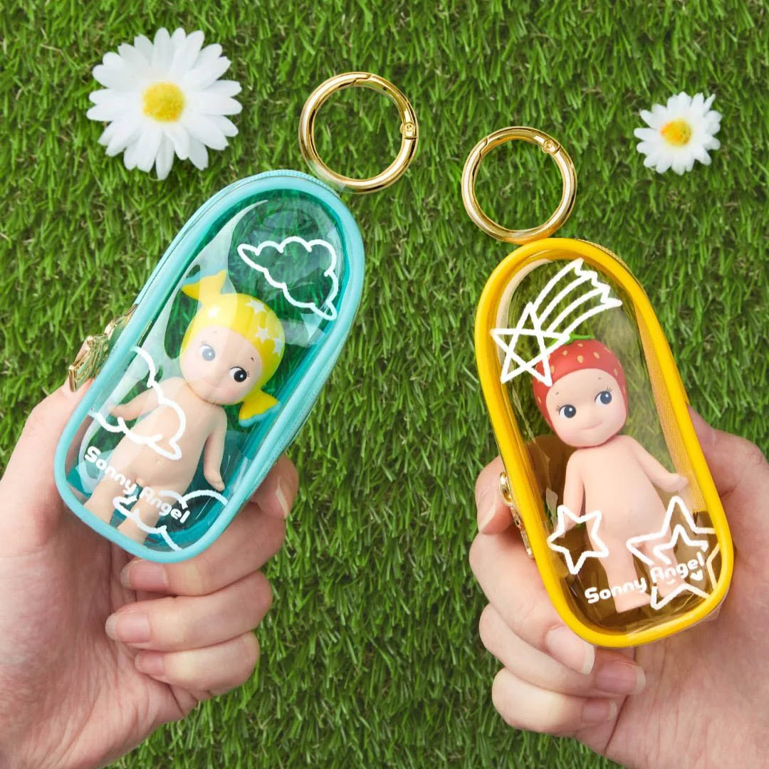 Zwei Hände halten je eine türkise und gelbe Schlüsselanhängertasche mit Sonny Angel-Figuren auf Rasen.