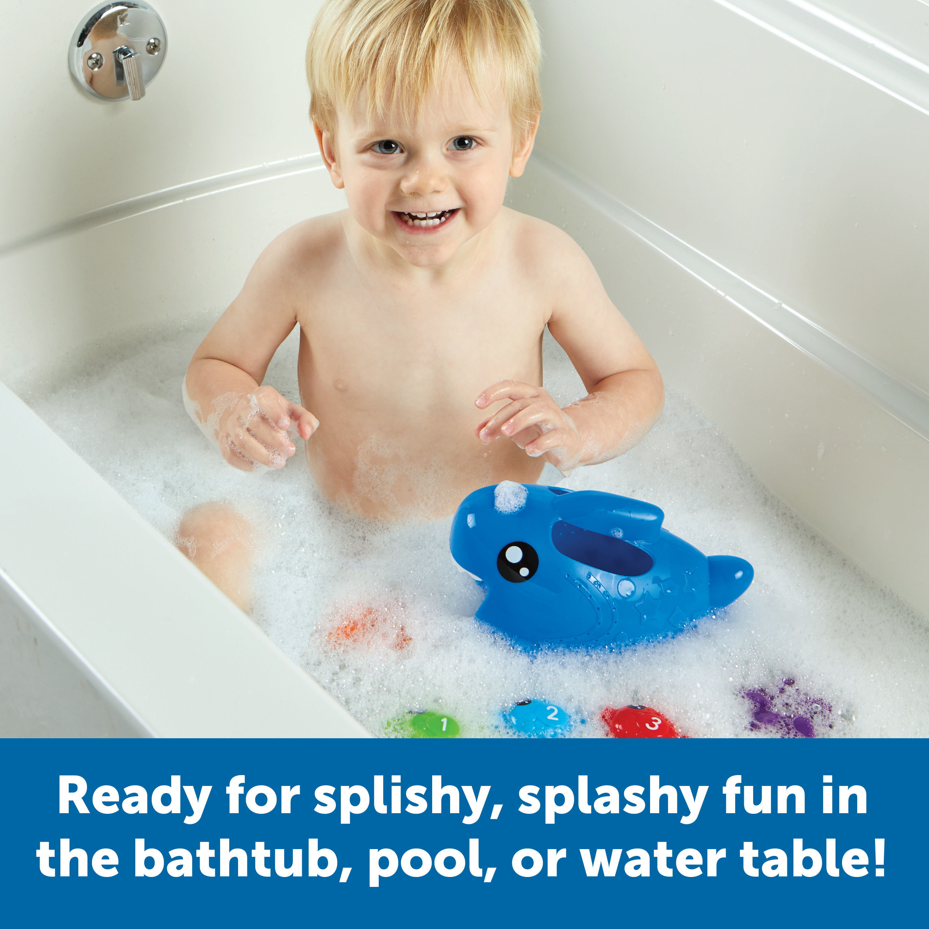 Kleinkind lächelt in einer Badewanne voller Schaum, mit einem blauen Wal-Spielzeug und bunten Fischen.