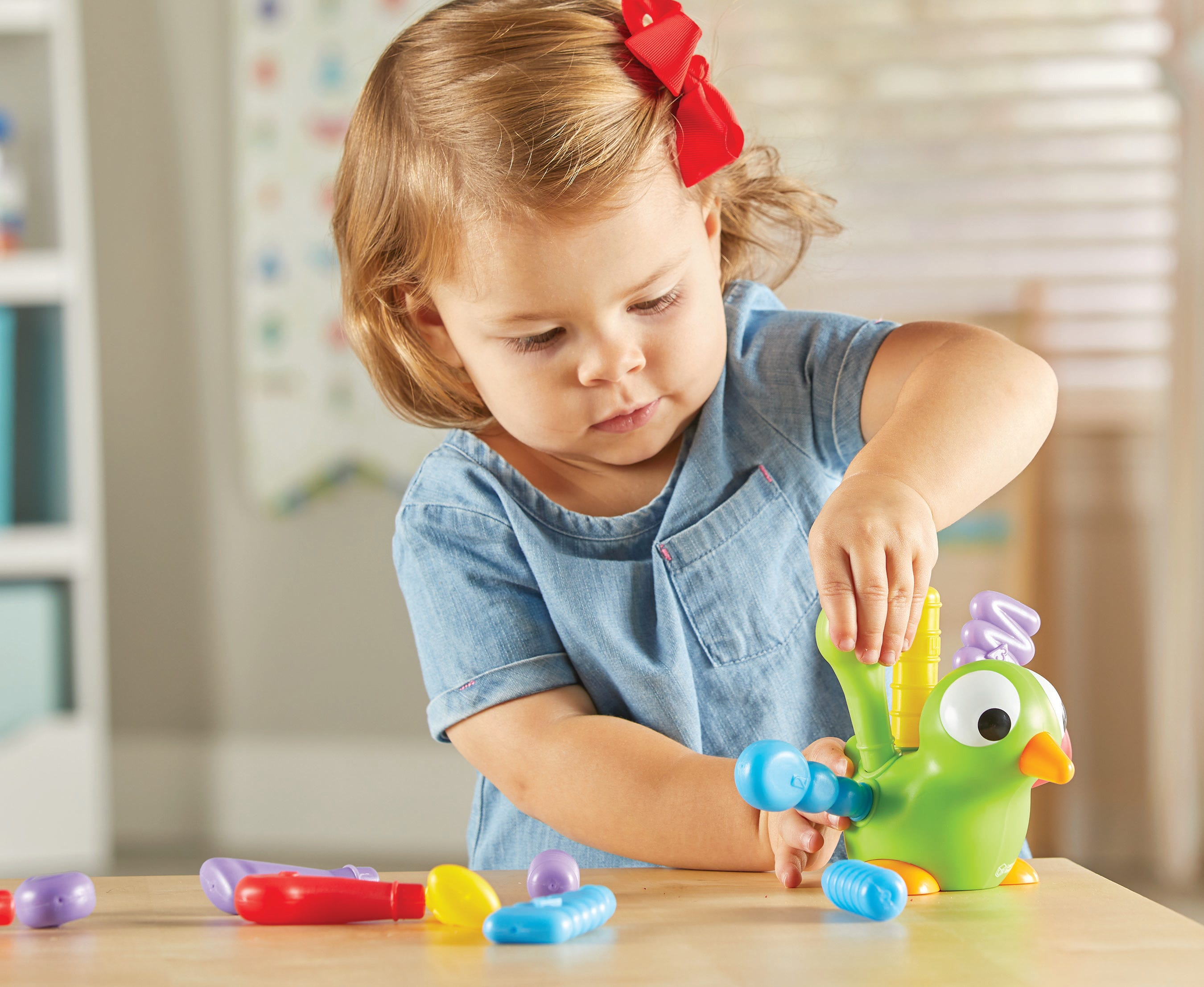 Kleines Mädchen in blauem Hemd und roter Schleife spielt mit grünem Spielzeugvogel am Tisch.