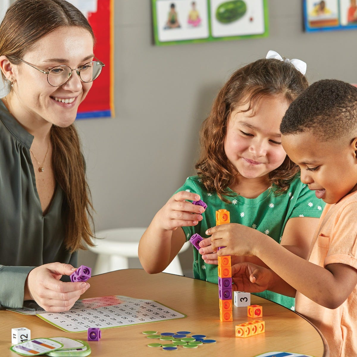 Eine Lehrerin sieht zu, wie ein Mädchen und ein Junge an einem Tisch einen Turm aus bunten Würfeln bauen.
