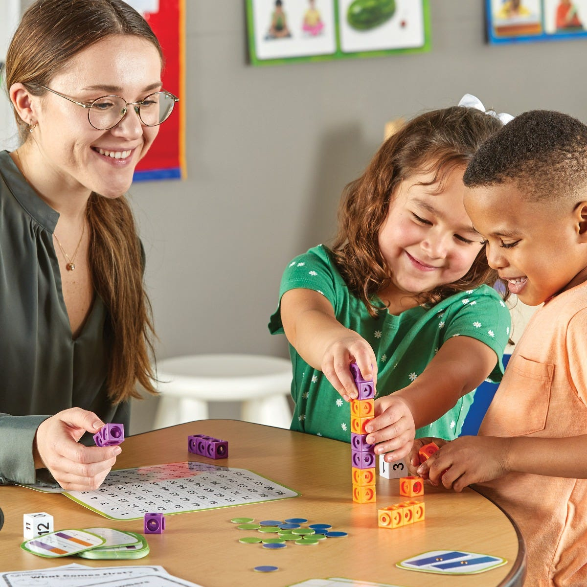 Eine Lehrerin und zwei Kinder bauen gemeinsam einen bunten Turm aus Plastikbausteinen an einem Tisch.