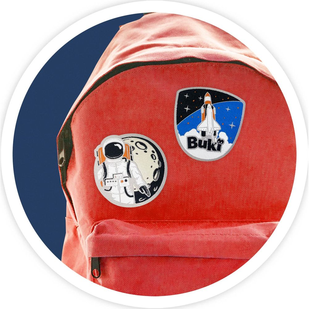 Ein roter Rucksack mit zwei Weltraum-Aufnähern: ein Astronaut vor dem Mond und eine Rakete mit dem Schriftzug Buki.