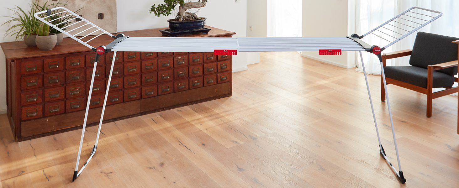 Ein weißer Wäscheständer steht auf einem Holzboden in einem hellen Raum mit Holzschrank und Stuhl im Hintergrund.