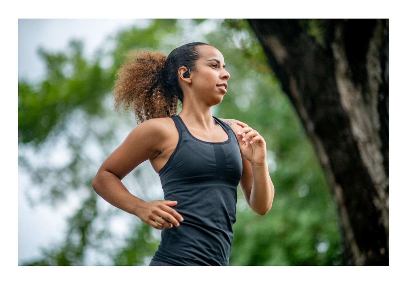Una mujer con una camiseta deportiva negra corre al aire libre, con árboles de fondo.