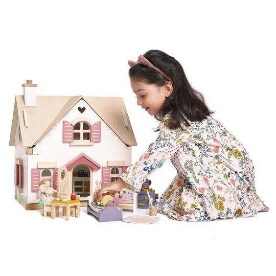 Ein Mädchen im Blumenkleid spielt auf dem Boden mit einem großen weißen und rosafarbenen Puppenhaus aus Holz.