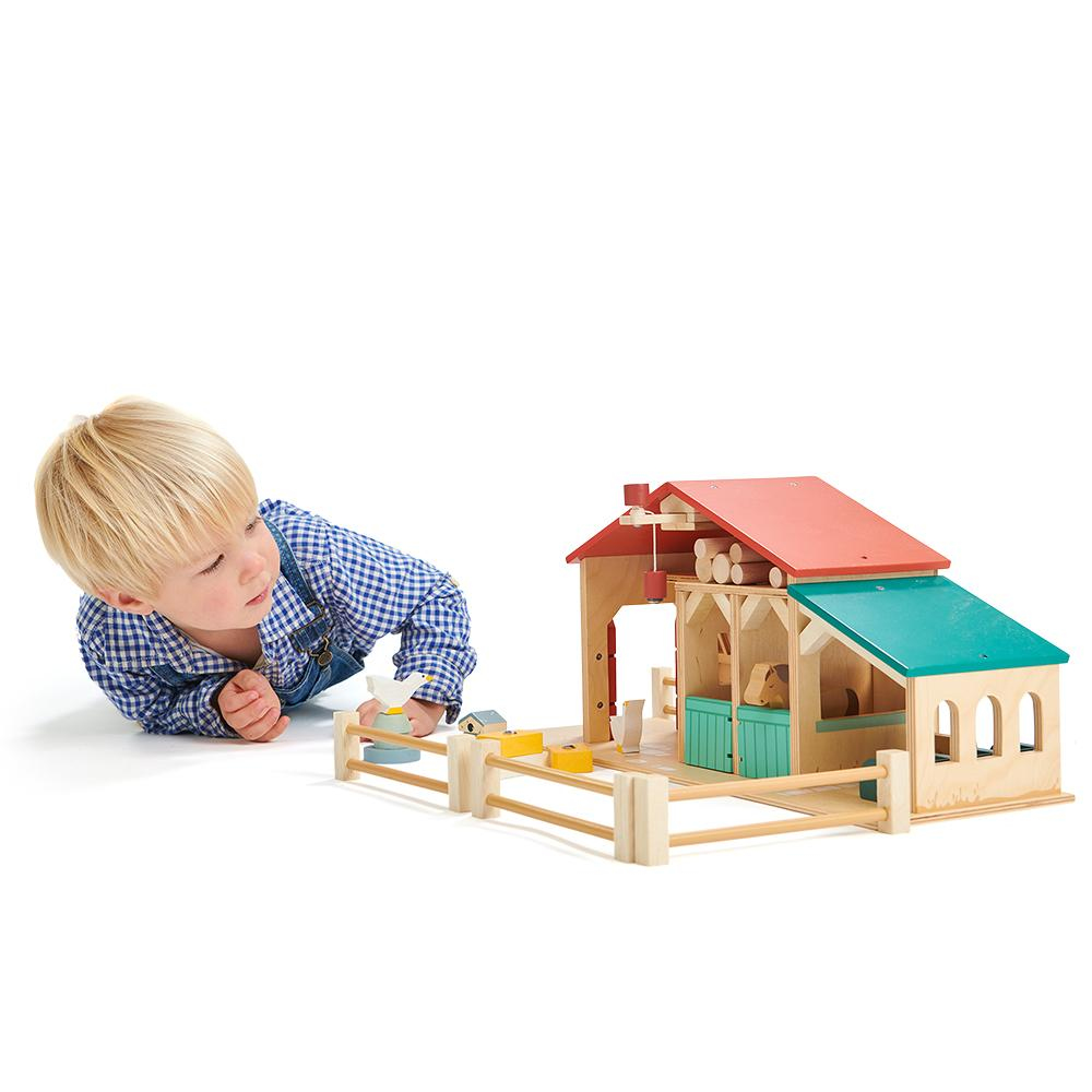Kleinkind in blauem Hemd spielt auf weißem Hintergrund mit einem bunten Holzbauernhof.