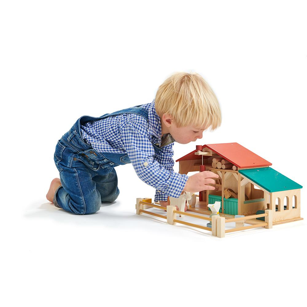 Kleiner Junge in Latzhose spielt auf weißem Boden mit einem Holzbauernhof.