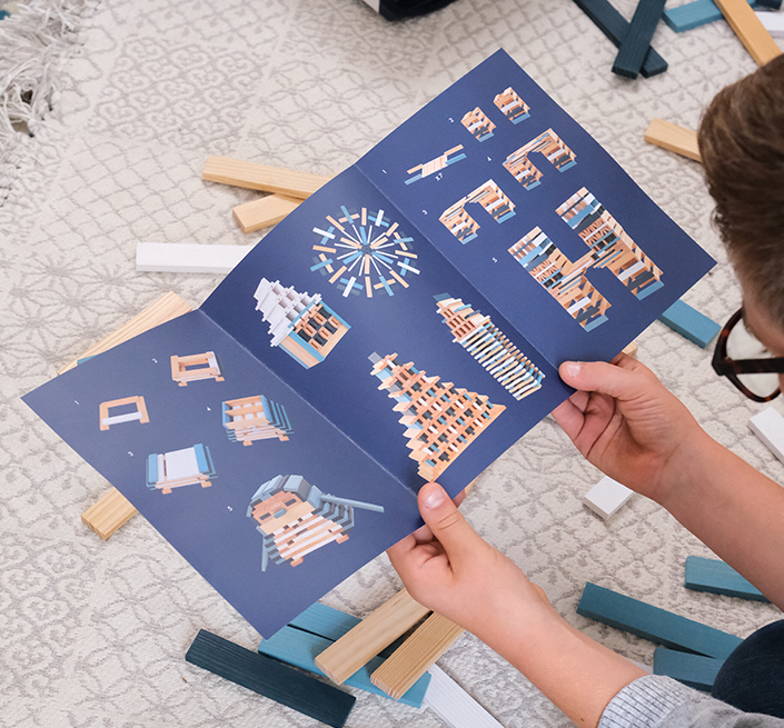 Ein Kind hält eine blaue Bauanleitung mit verschiedenen Holzblockmodellen in den Händen.