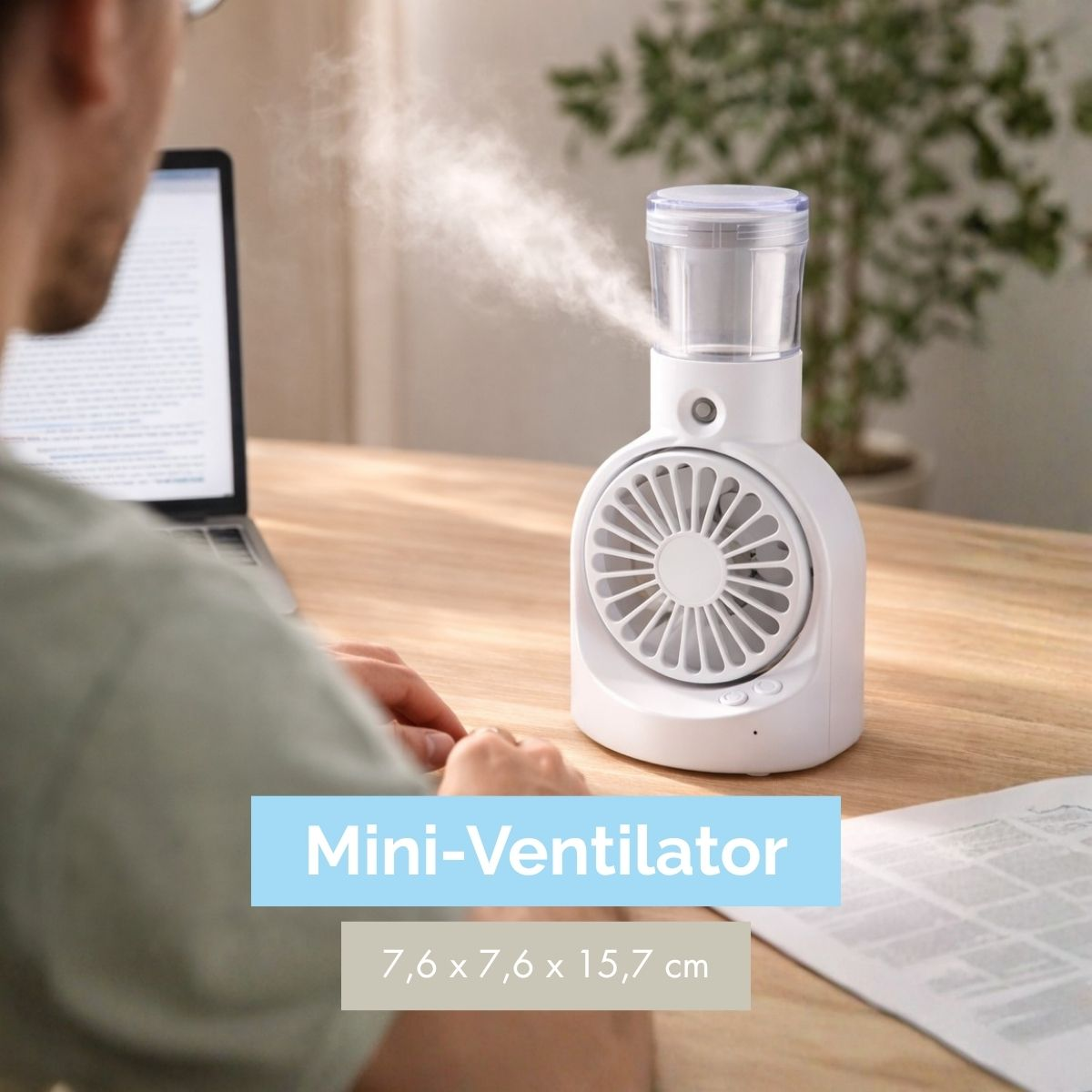 Ein weißer Mini-Ventilator mit Wassertank sprüht Nebel auf einem Holztisch, neben einem Laptop.