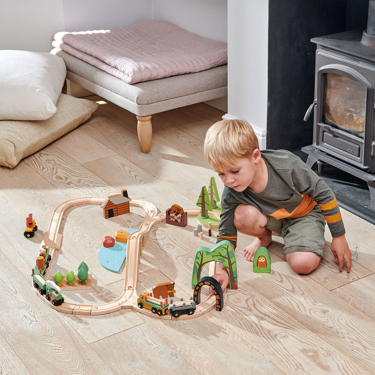 Ein kleiner Junge spielt auf Holzboden mit einer Holzeisenbahn neben Kamin und Kissen.