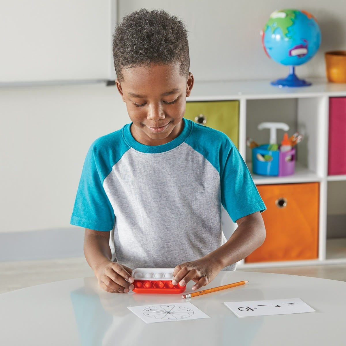 Ein Junge in einem blau-grauen T-Shirt sitzt am Tisch und spielt mit einem roten Push-Pop-Spielzeug.