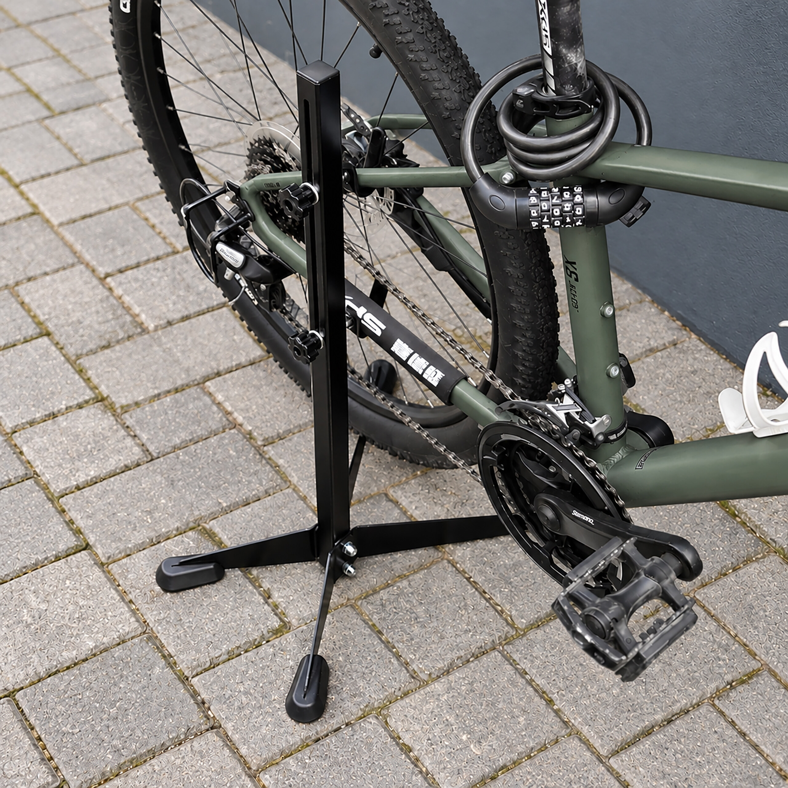 Ein grünes Mountainbike steht auf einem Ständer auf Pflastersteinen, gesichert mit einem Kabelschloss.