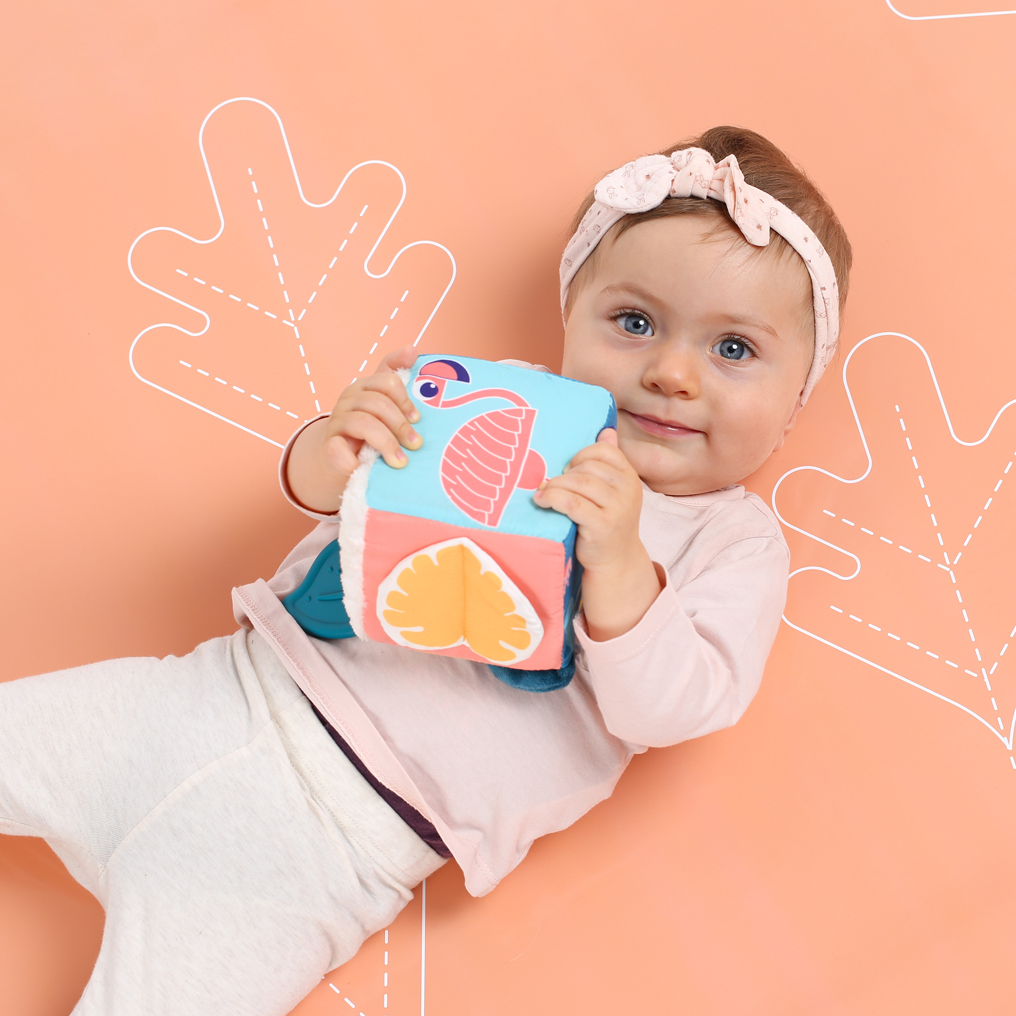 Ein lächelndes Baby mit einem Haarband liegt auf einer rosa Decke und hält einen bunten Spielzeugwürfel.