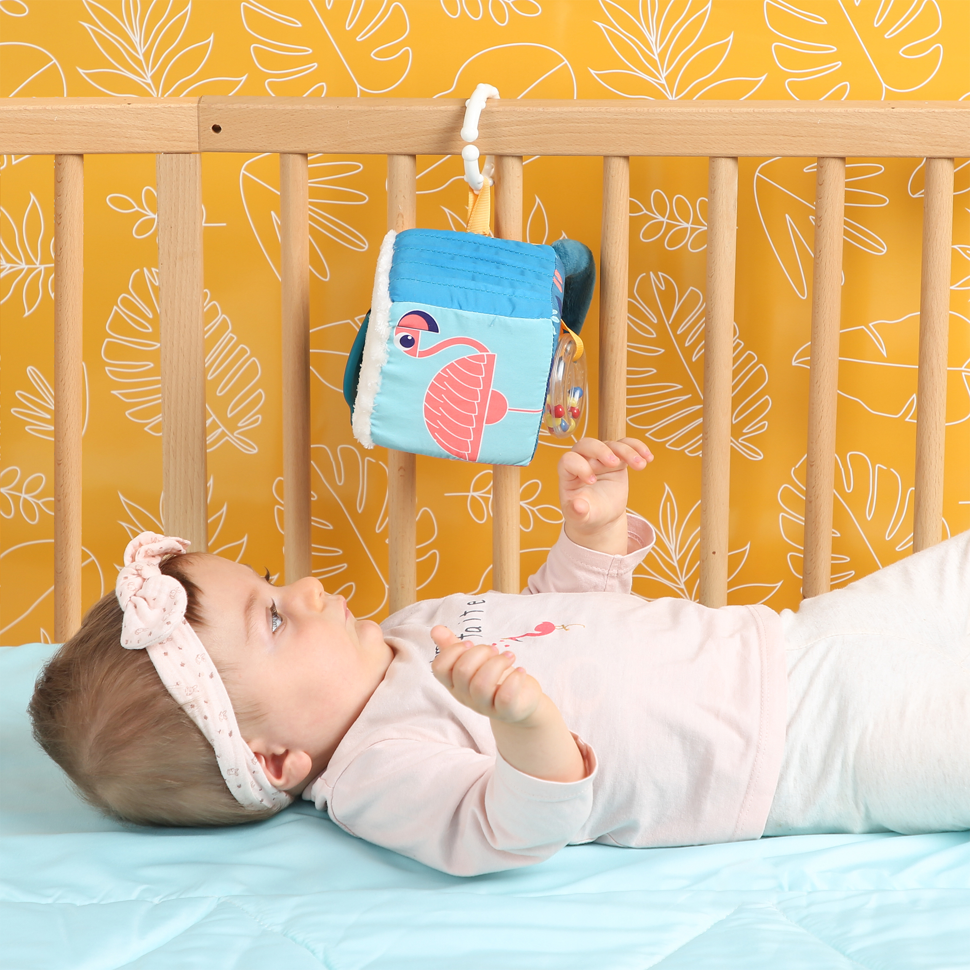Ein Baby in Rosa liegt im Bett und schaut zu einem hängenden Stoffwürfel mit Flamingomotiv.