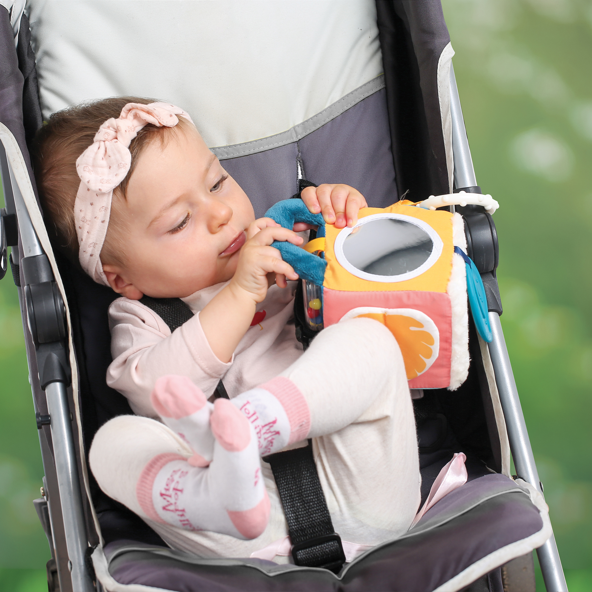 Kleines Mädchen mit rosa Haarschleife und Socken sitzt im Kinderwagen und spielt mit einem bunten Spielzeug.