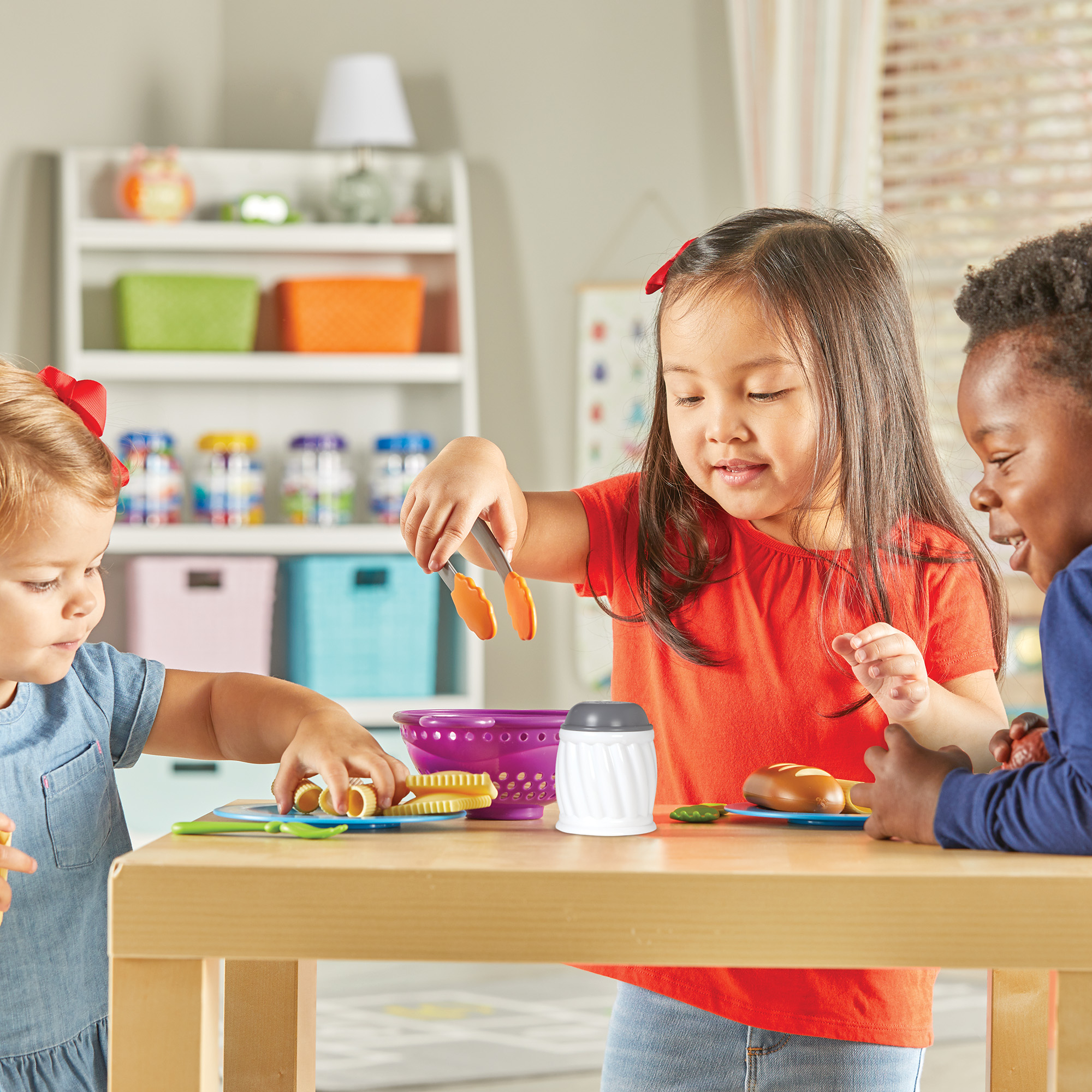 Drei Kinder spielen an einem Holztisch mit bunten Spielzeuglebensmitteln und Zubehör in einem hellen Raum.