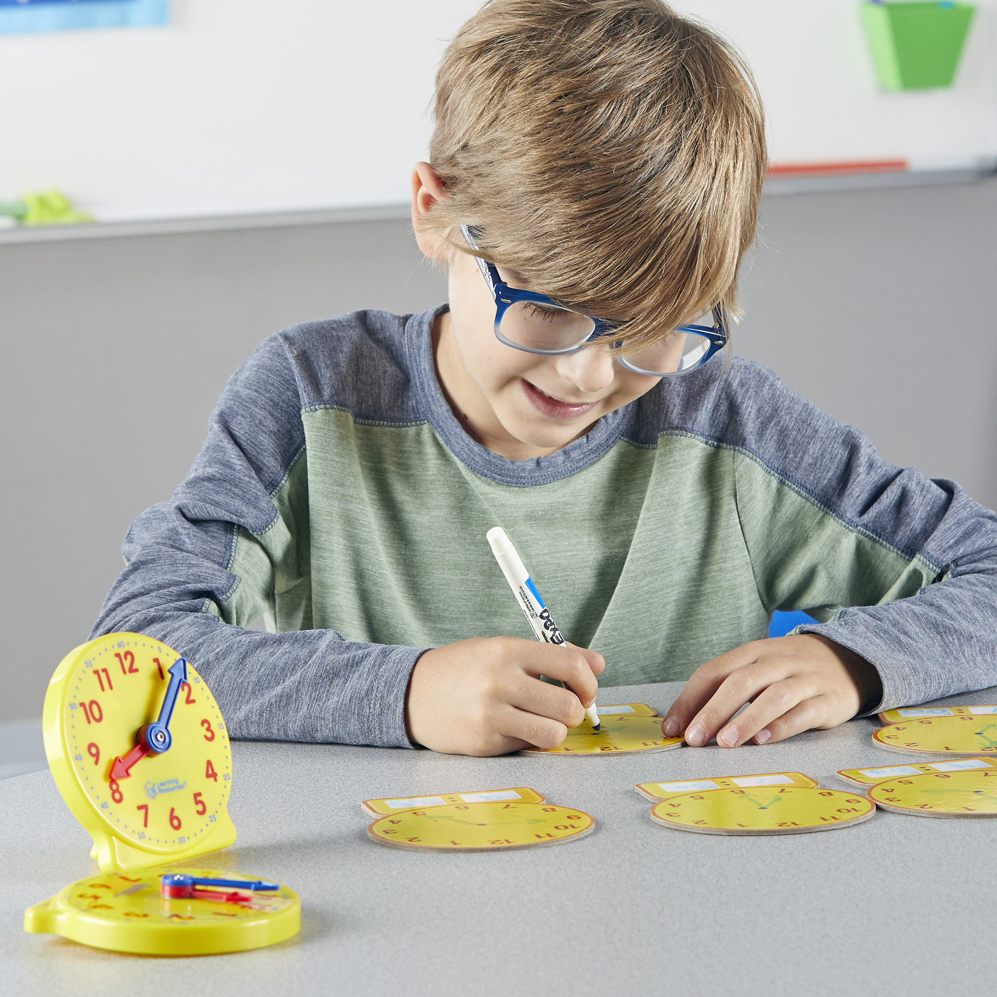 Ein Junge mit Brille malt mit einem Stift auf ein gelbes Lernzifferblatt am Tisch.