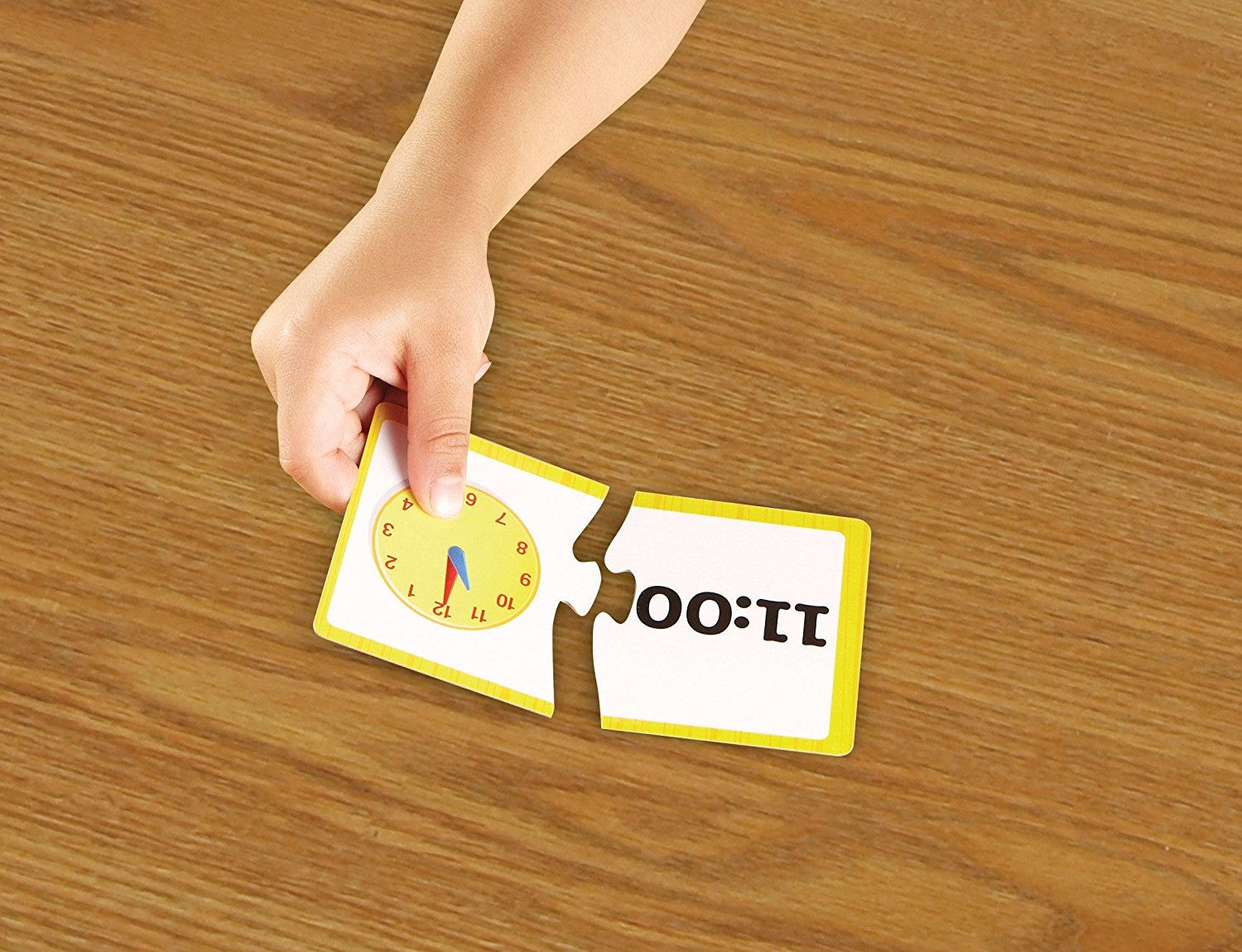 Eine Kinderhand verbindet zwei Puzzleteile auf einem Holztisch, eins mit Uhr um 11:00 und das andere mit Text.