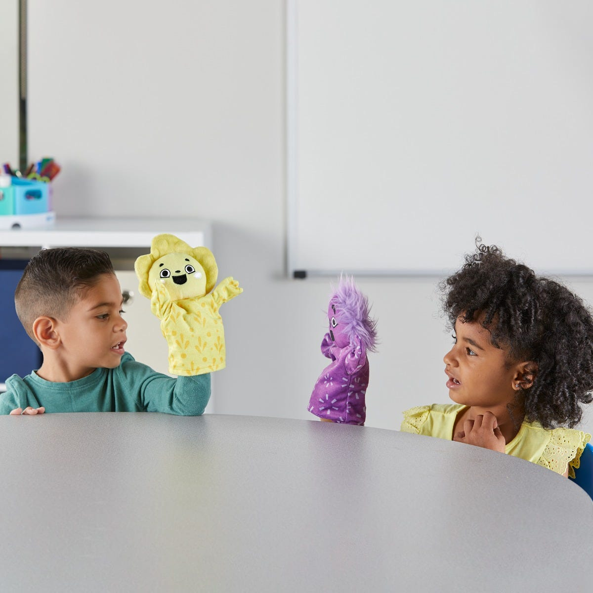 Zwei Kinder spielen an einem runden Tisch mit Handpuppen: ein Junge mit einer gelben und ein Mädchen mit einer lila Handpuppe.