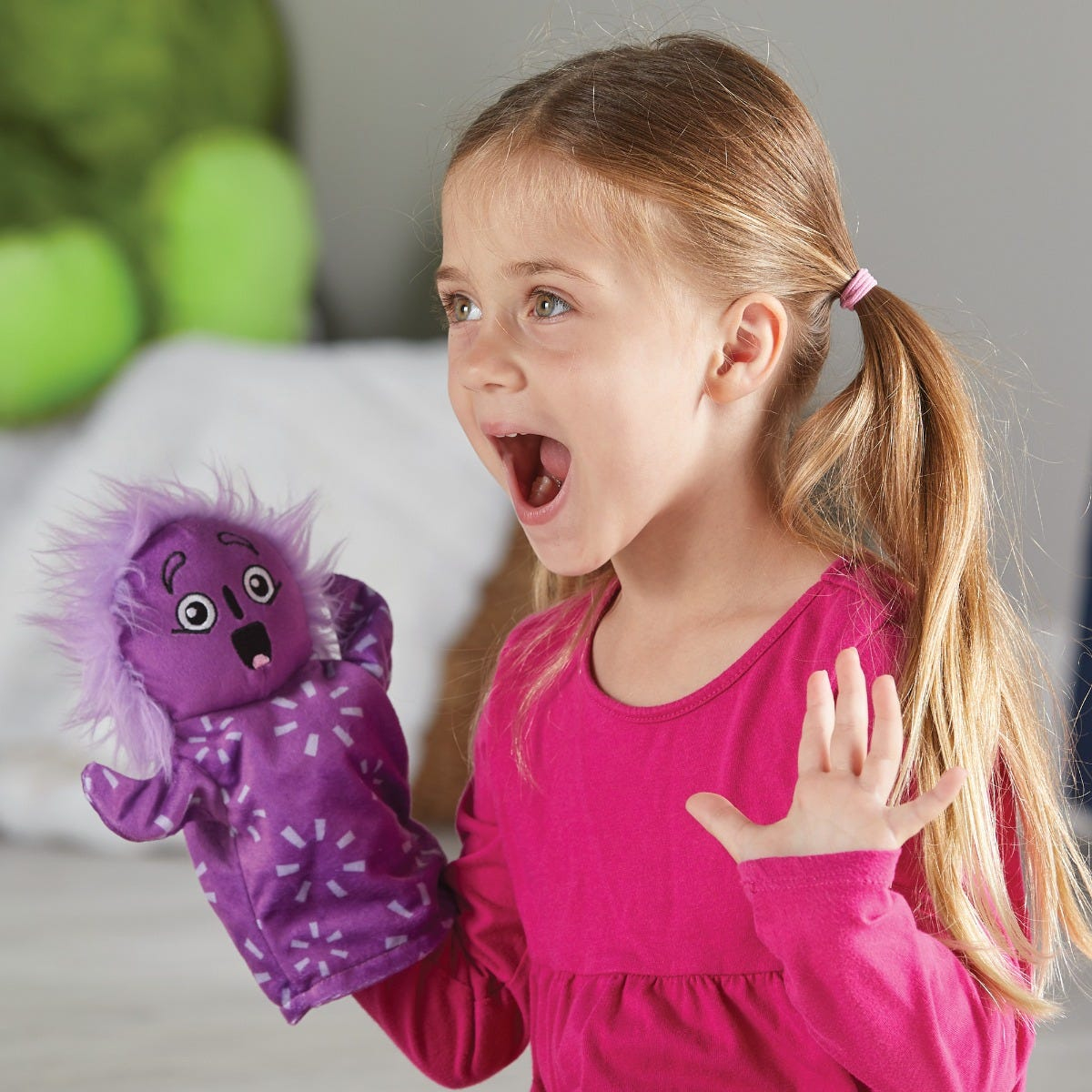 Ein blondes Mädchen in Rosa hält eine violette Handpuppe und ruft fröhlich.