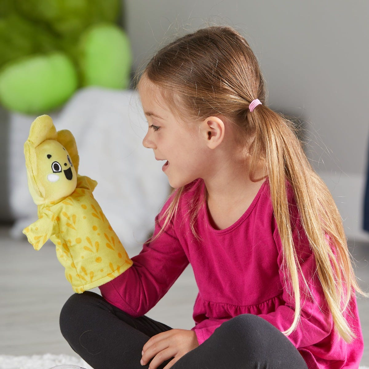 Ein junges Mädchen mit langem blonden Haar sitzt und spielt lächelnd mit einer gelben Handpuppe.