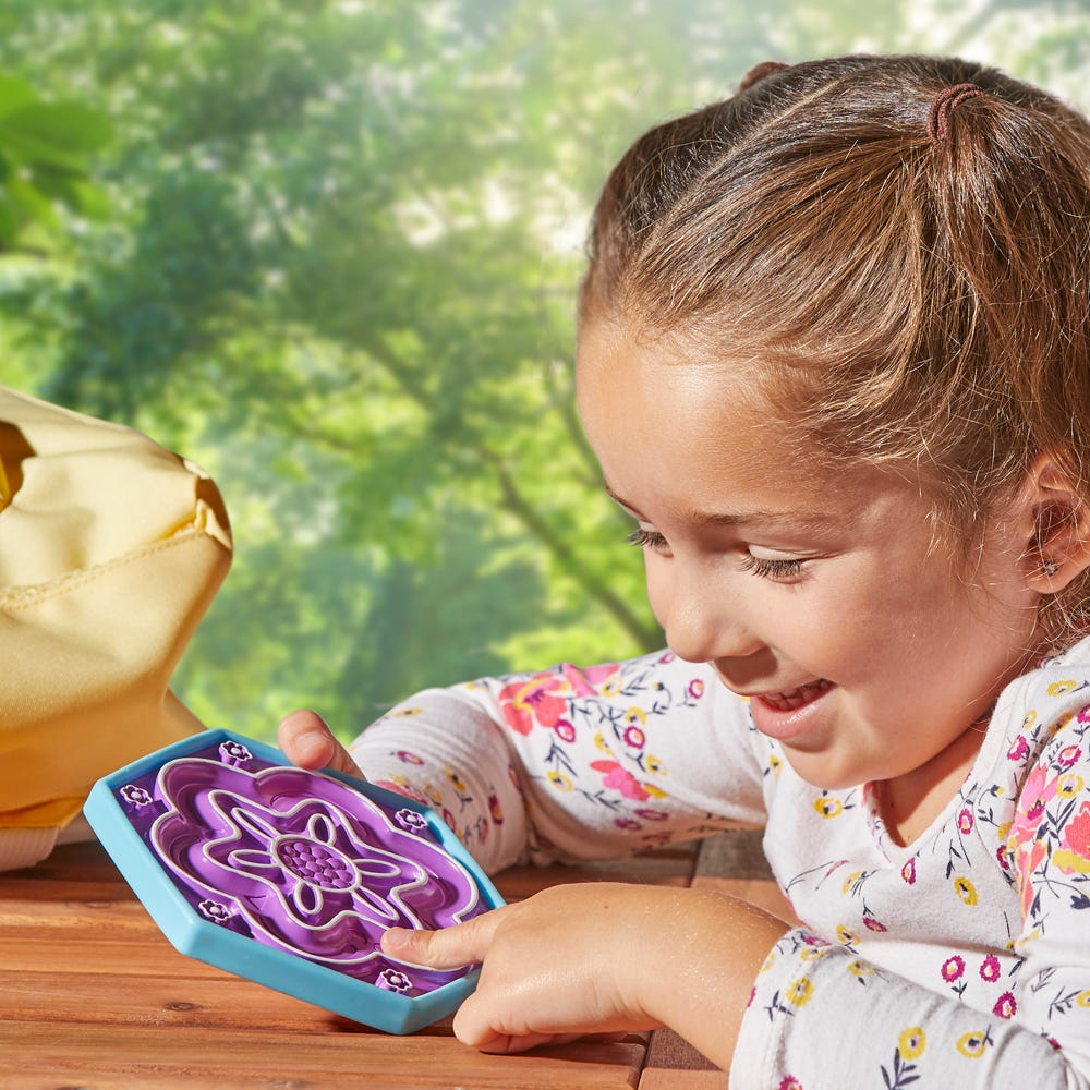 Ein junges Mädchen mit zwei Zöpfen spielt draußen an einem Holztisch mit einem blau-violetten Geschicklichkeitsspiel.