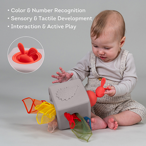 Ein Baby spielt mit einem grauen sensorischen Würfel, der bunte Tücher und eine rote Hasenfigur enthält.