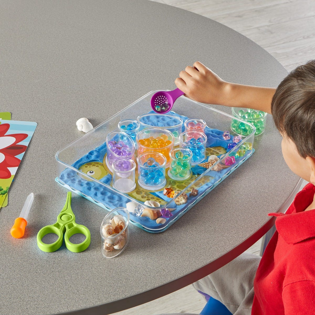 Ein Kind spielt mit bunten Wasserperlen und Muscheln in einer transparenten Schale.