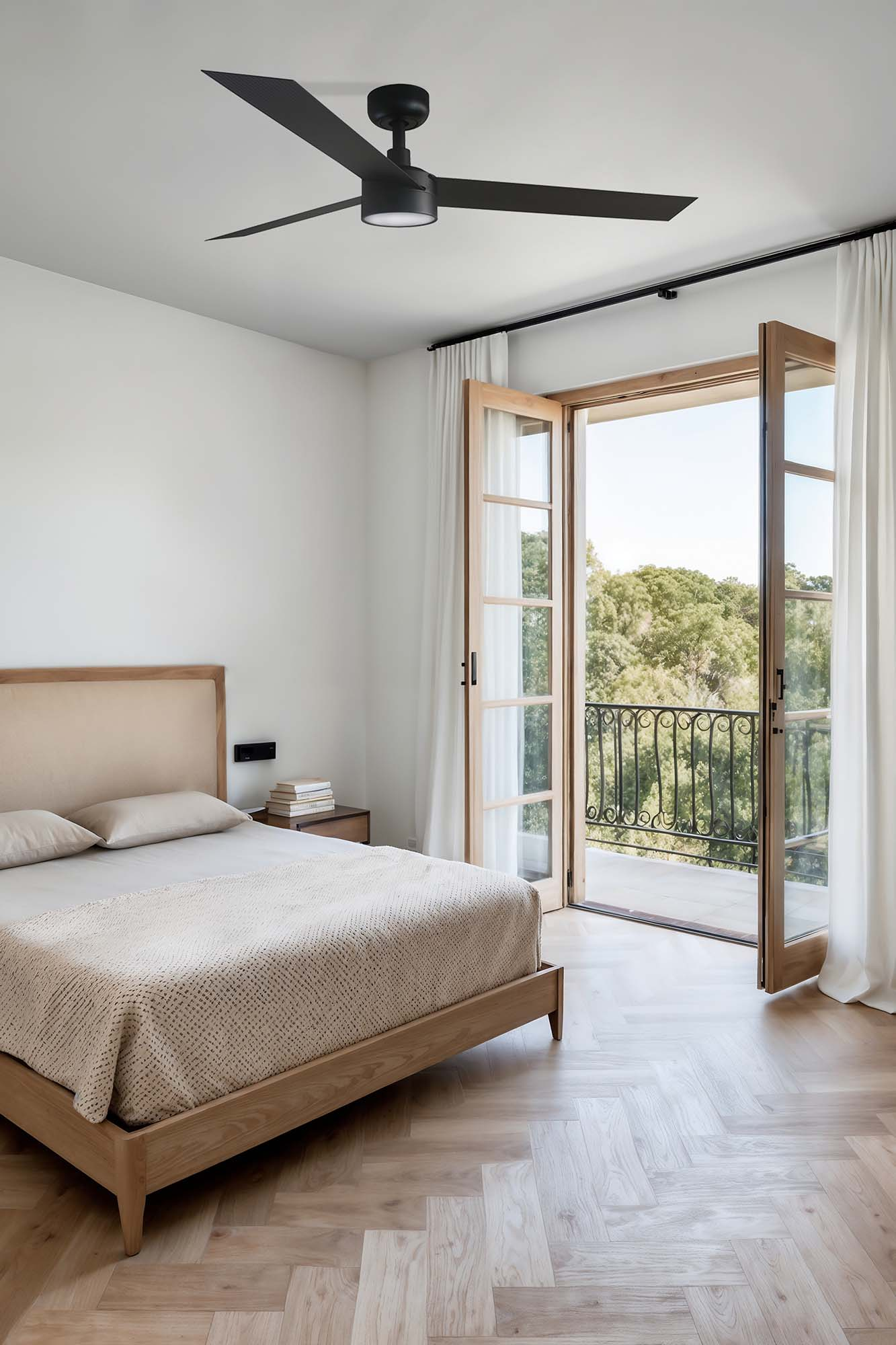 Ein helles Schlafzimmer mit Holzbett, Fischgrätparkett und offenem Balkon mit Blick auf Bäume.