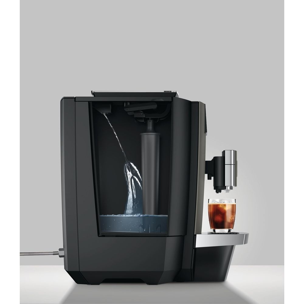 Eine schwarze Kaffeemaschine mit sichtbarem Wassertank füllt ein Glas mit Kaffee auf einer grauen Oberfläche.